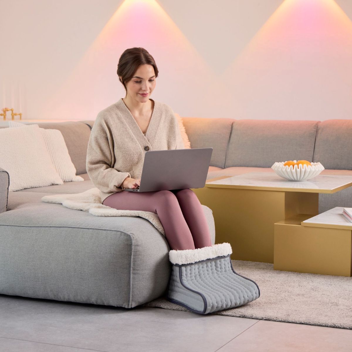 A woman sits on a sofa working on a laptop while wearing a grey electric foot warmer in a modern living room.